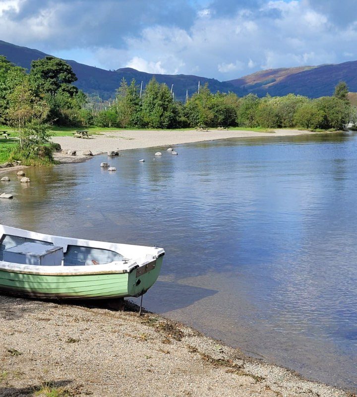 a green and white boat on the bank of a blue lake with trees and mountains in the distance