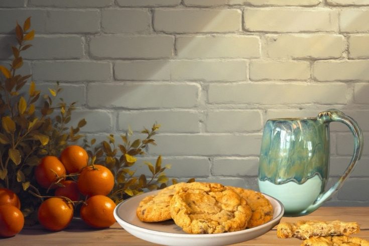 a white brick wall background with a green mu and a plate of chocolate chop cookies. To the left is some greenery and orange tomatoes
