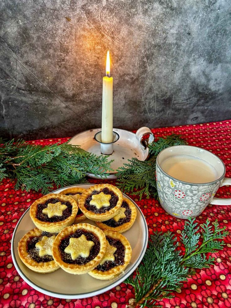A red tablecloth with a plate of mince pies, each mince pie has a pstry star on top. to the right is a cup of hot chocolate and behind is a white candel. surrounding the plate are some conifer branches