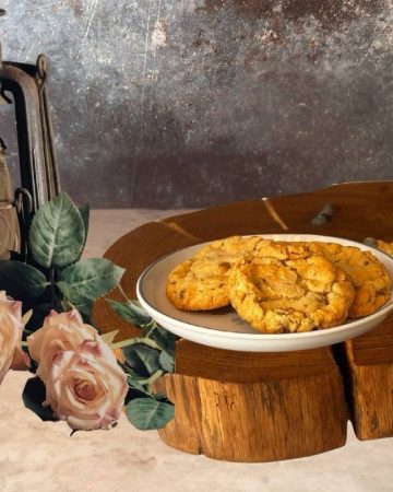 a slice of wood with a plate of cookies and a few chocolate chips and a handmade mug on. to the left is an oil lamp with some pink roses in front